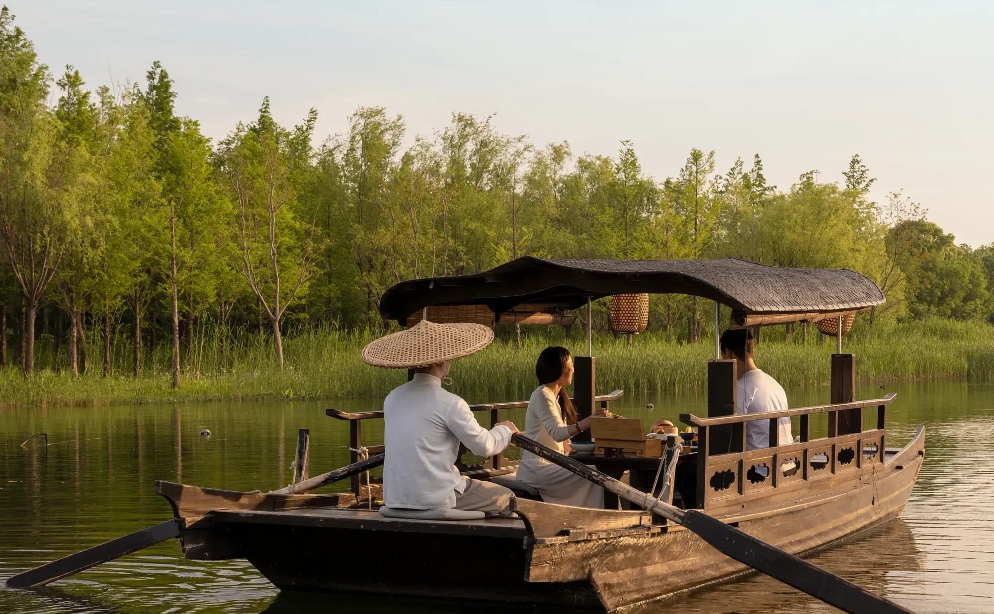 A wooden boat with a thatched canopy carrying two guests across the water at low sun, rowed by a boatman in a conical straw hat
