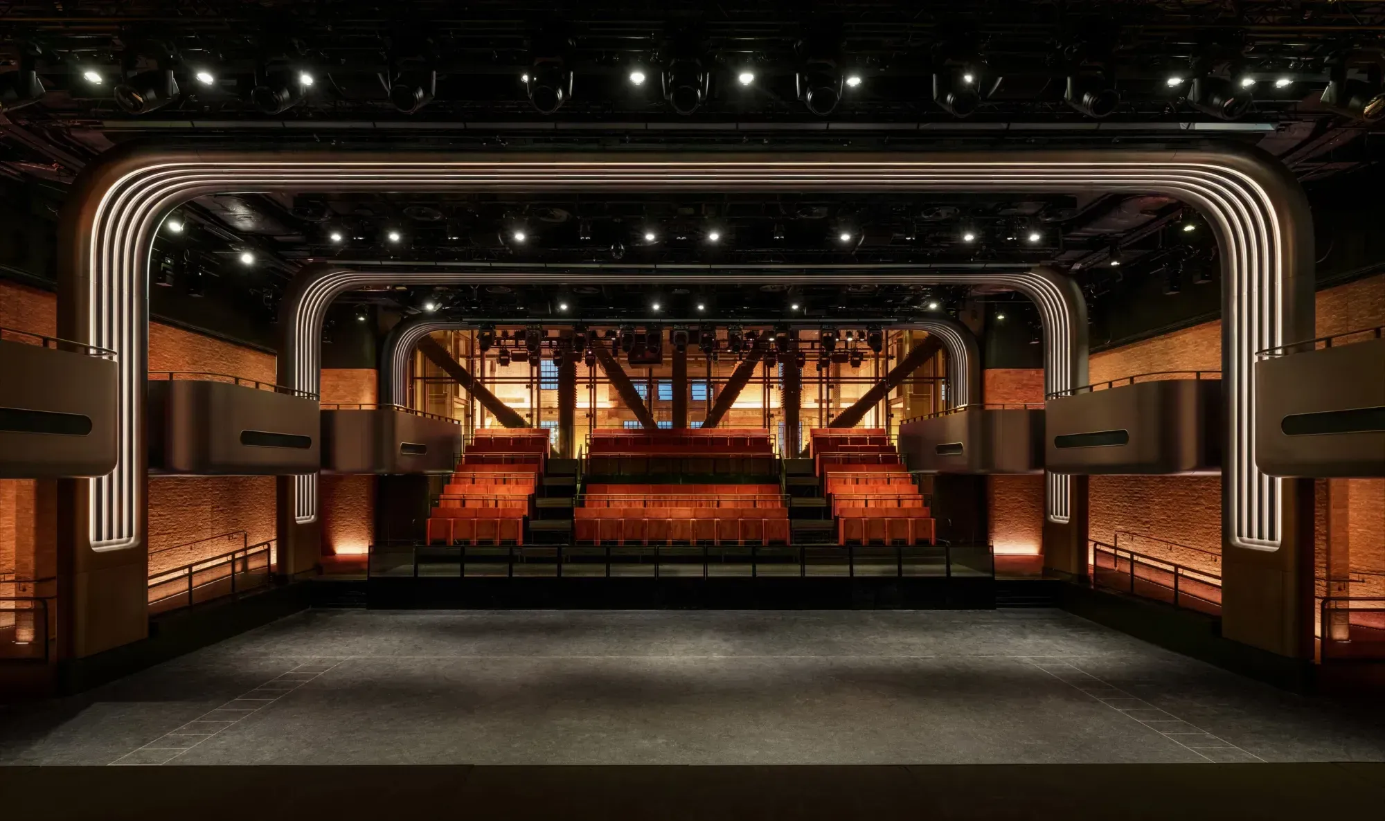 The auditorium of Apple Music Hall seen from the stage, with banked rows of terracotta seats framed by nested arches of warm strip light, exposed brick walls, bronze balcony boxes and a glass wall behind the seating showing the steel structure of Battersea Power Station