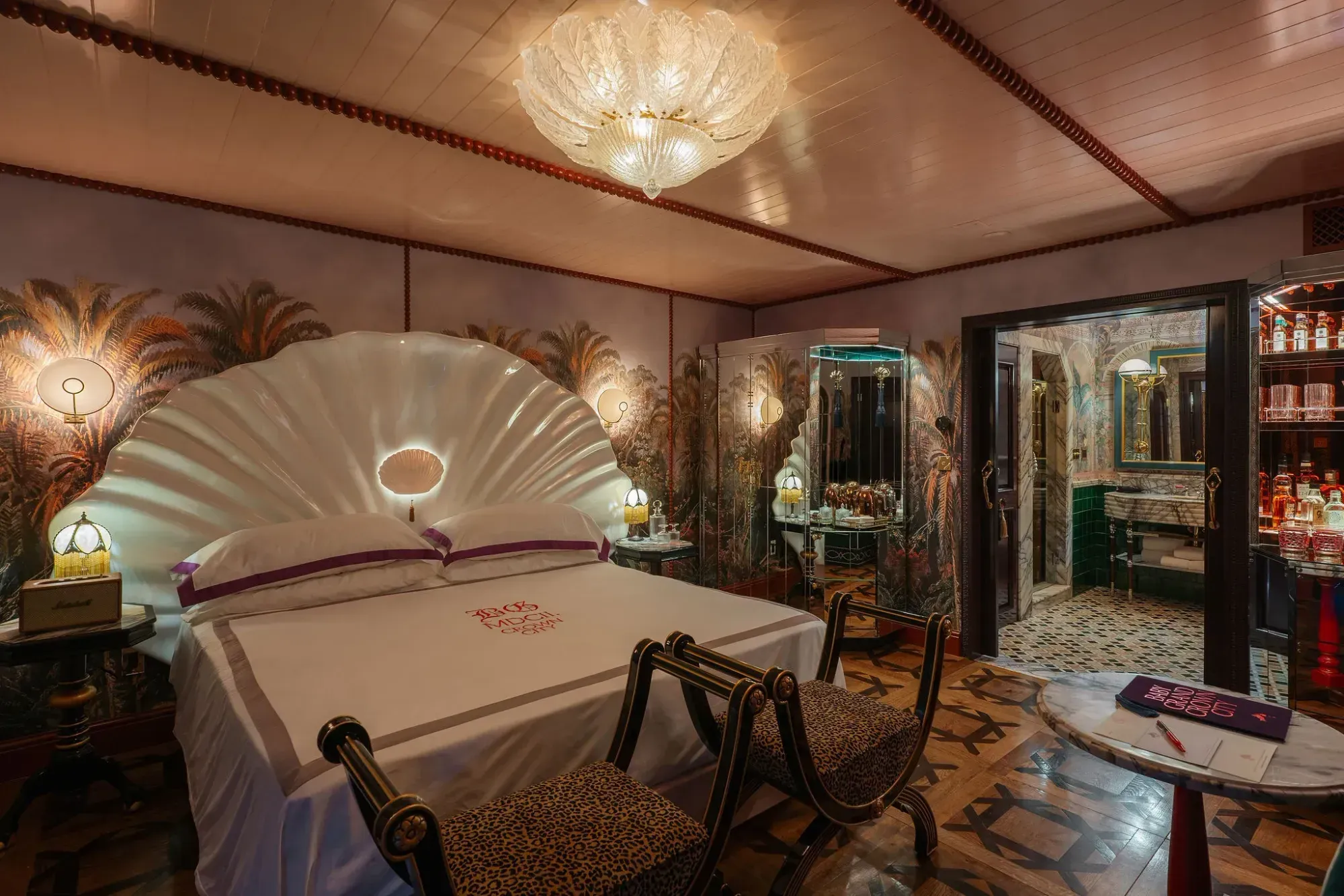 A hotel bedroom with a tall iridescent clamshell headboard, a painted palm mural on the walls, a mirrored bar cabinet lit red, a leopard-print rug and a shell-shaped ceiling light