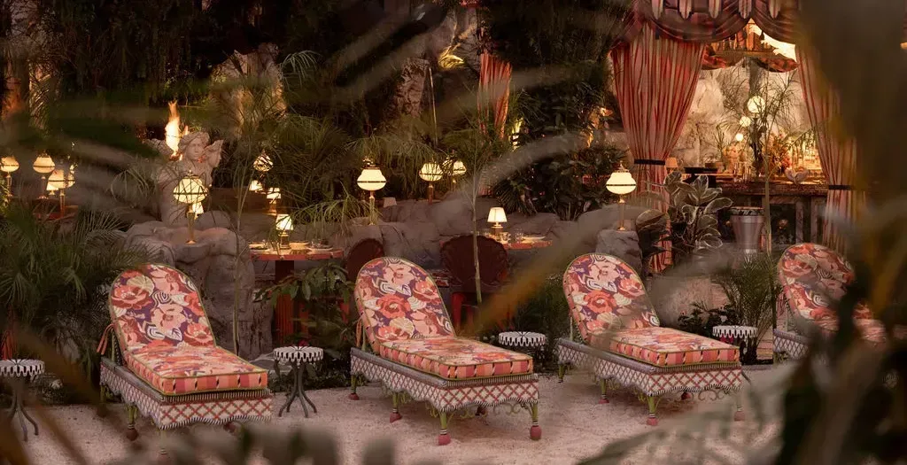 Four floral-upholstered chaise longues on pale sand under hanging greenery and a striped canvas canopy, with small shaded lamps glowing on rocks and tables behind them at dusk