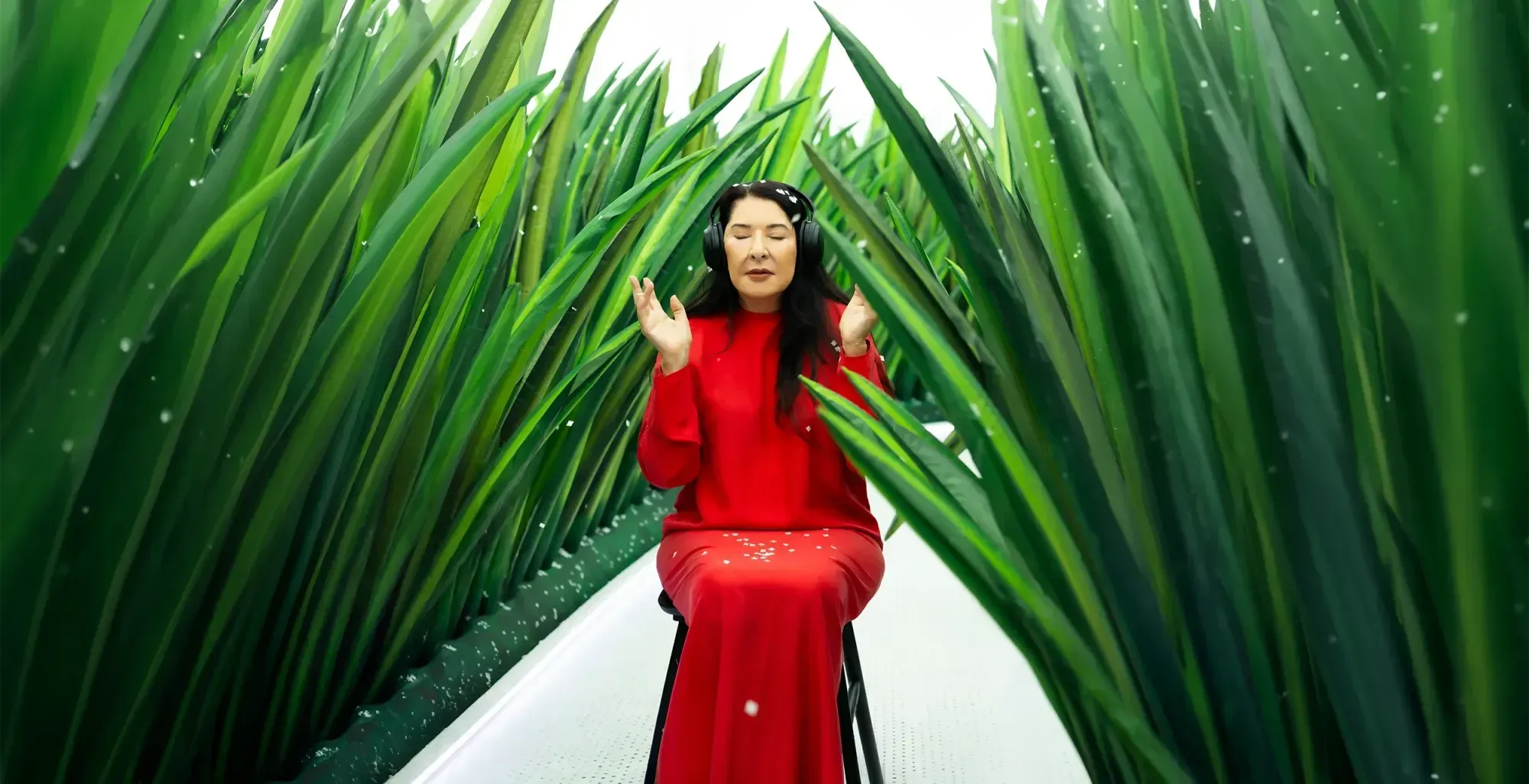 Marina Abramović in a red dress and headphones sits on a white path between walls of tall inflatable grass as paper snow falls, in Snowy / Windy / Spring on the Planet Z