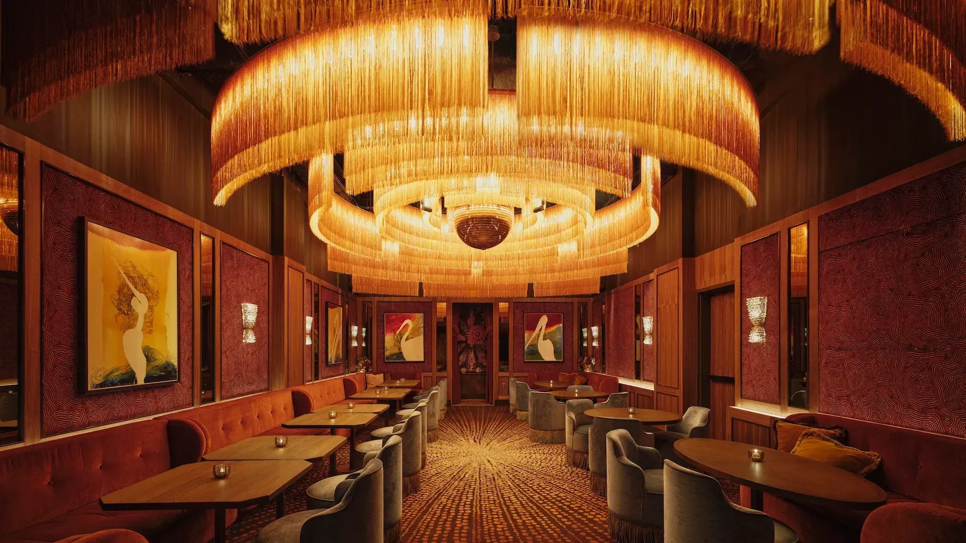 A long room under a tiered, fringed ring chandelier, with rust velvet banquettes and grey fringed armchairs along both walls, red velvet panels hung with paintings of a white bird, and a patterned carpet down the centre aisle