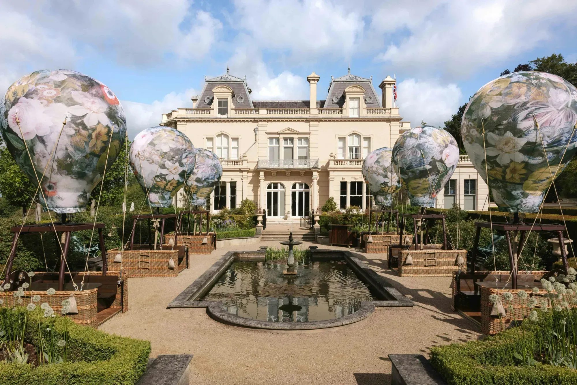 Six hot air balloons printed with a pale floral pattern stand in two rows either side of a rectangular reflecting pool, with the chateau front of Beaverbrook's main house behind.