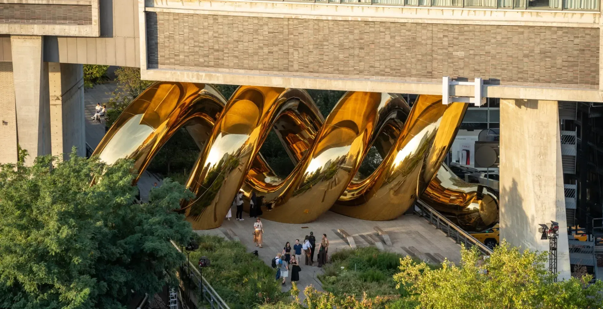 Seen from above in low evening sun, a run of giant mirror-polished gold coils fills the covered stretch of the High Line beneath The Standard, with people gathered on the deck at their base