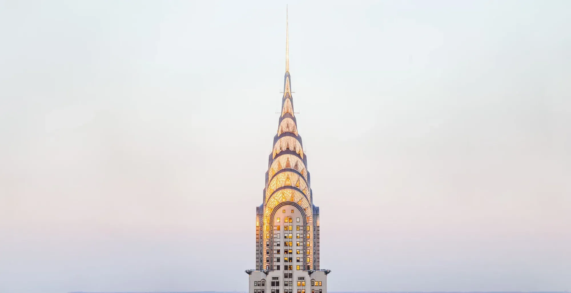 The Chrysler Building's crown and spire at dusk, the stainless-steel arches lit gold against a pale sky