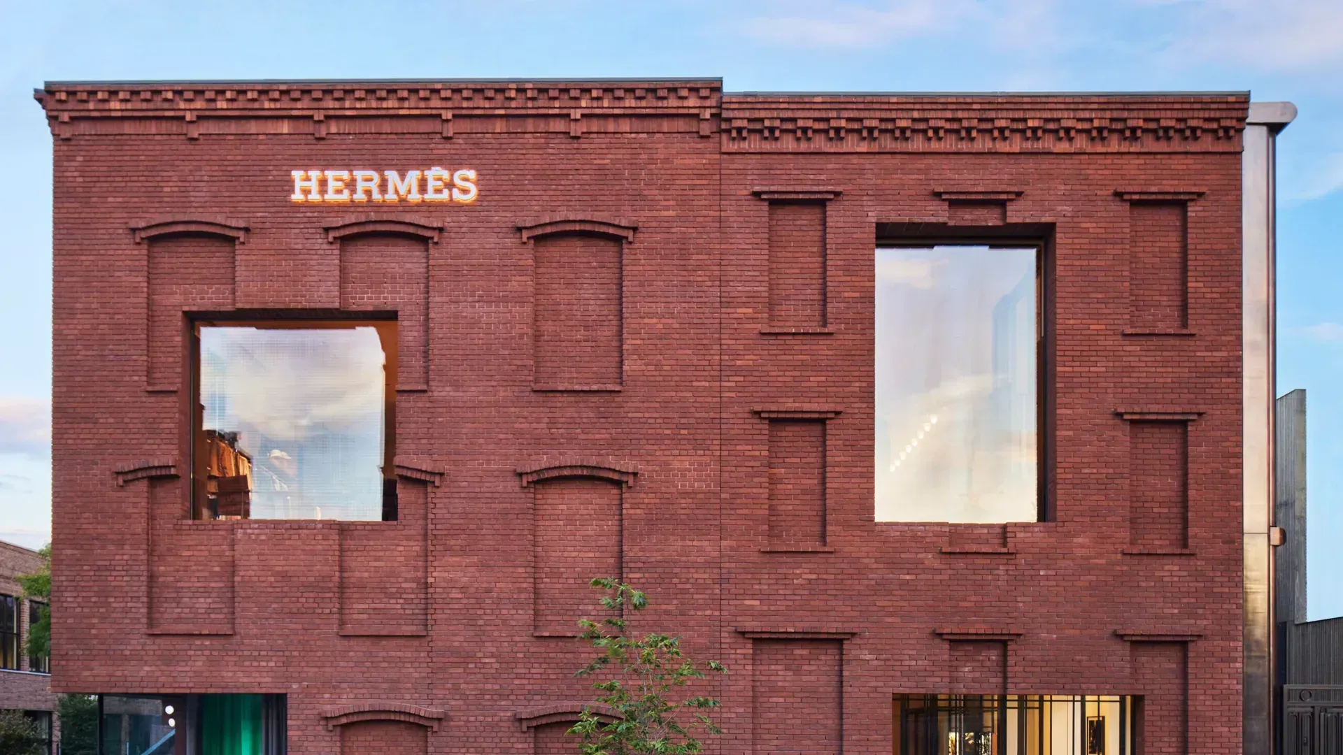 Two-storey red brick building with bricked-in arched windows, two large glazed openings on the upper floor, illuminated Hermès lettering and colorful window displays at street level, a young tree on the sidewalk