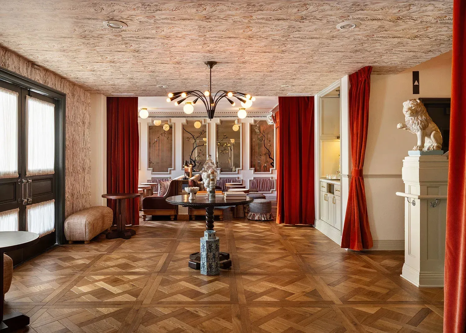 Hotel lobby with marquetry parquet floors, a ceiling papered in a mottled stone print, a black chandelier with globe bulbs, red velvet curtains and a white plaster lion on a plinth