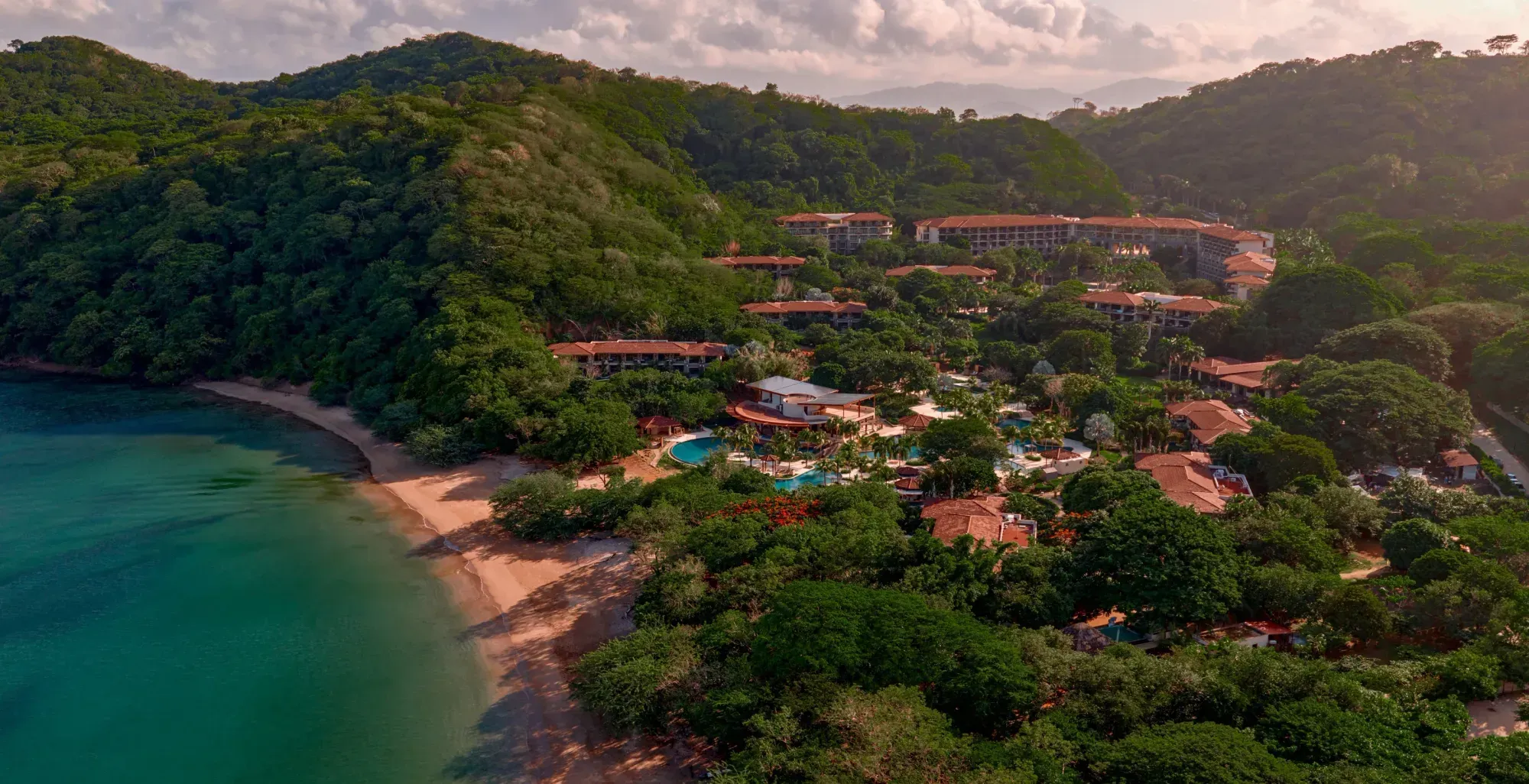 Aerial view of a curved sandy beach and turquoise bay, with terracotta-roofed resort buildings and pools set into forested hills