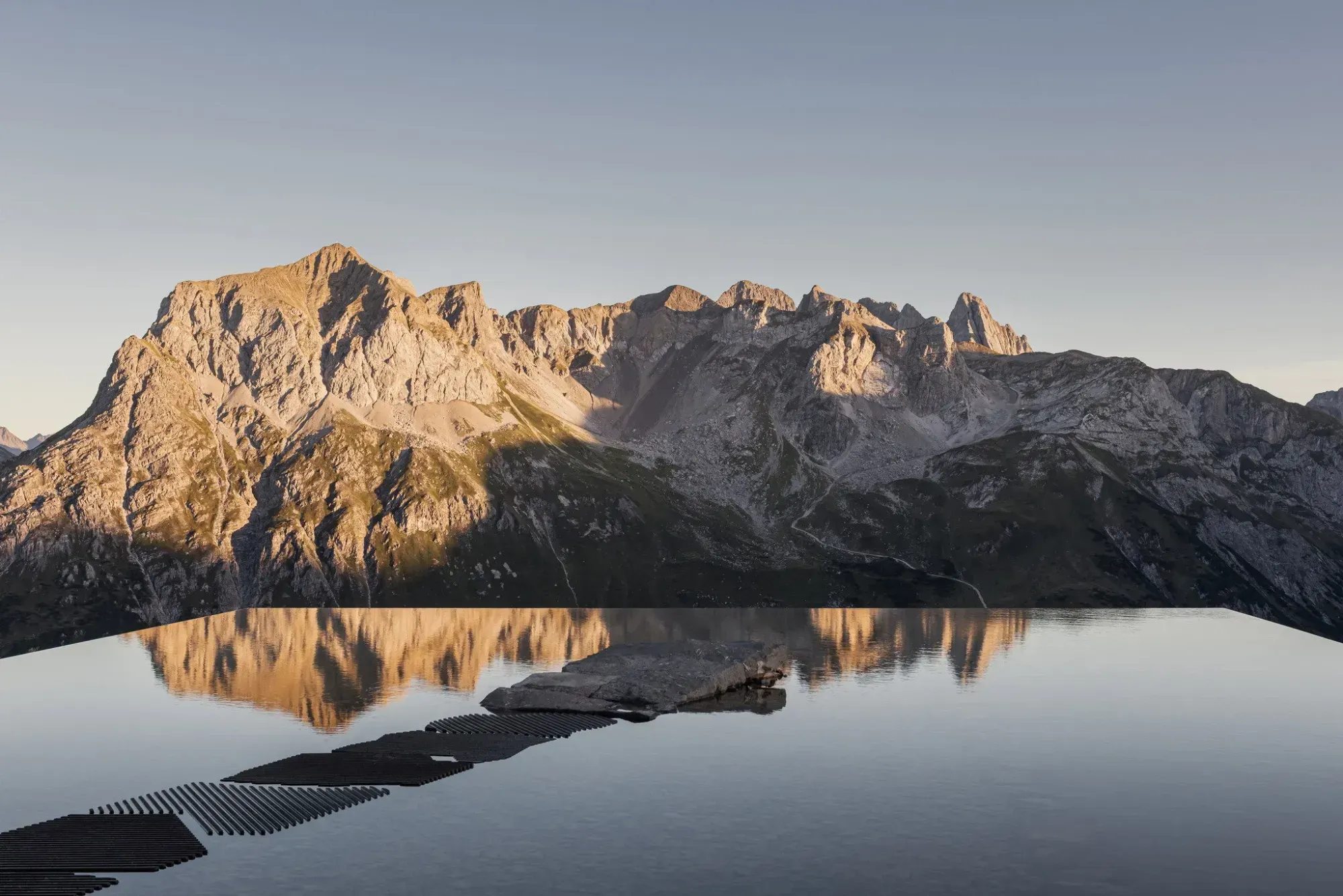 A shallow rooftop pool reflecting a mountain ridge lit gold by low sun, with a line of dark ribbed stepping stones leading across the water to a flat rock