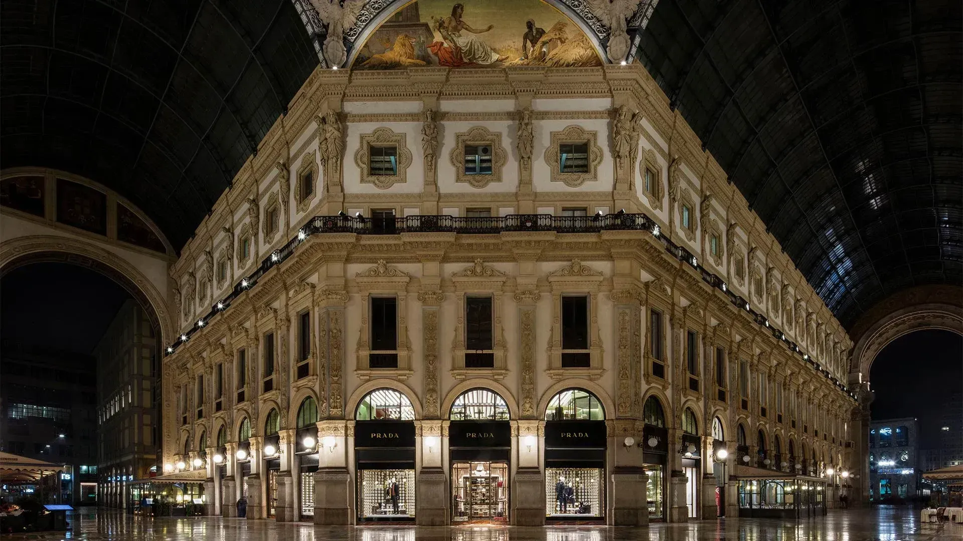 The cream stone facade of a Galleria Vittorio Emanuele II palazzo at night under the glass vault, with three arched shop windows lettered PRADA at street level and a painted lunette above