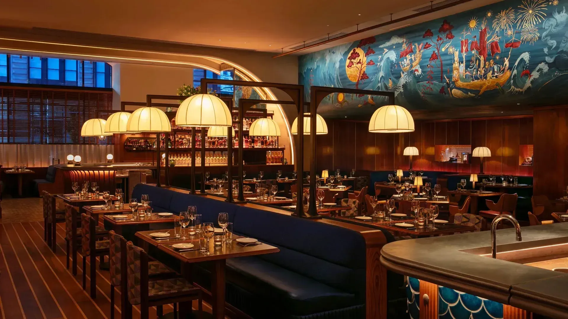 An empty dining room with wood-panelled walls, a long blue banquette, pleated lamp shades on dark metal frames, a raw bar faced in blue scale tiles, and a painted mural of waves, fireworks and a boat of figures along the upper wall