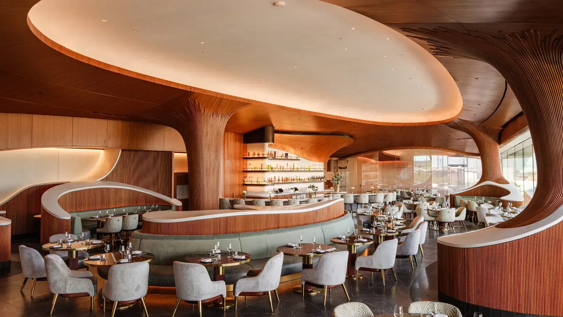 A dining room of curved cherry wood under a lit oval ceiling, with a tree-like cherry column, a sage-green banquette ringing a central island, grey chairs at round tables and a bar lined with bottles behind