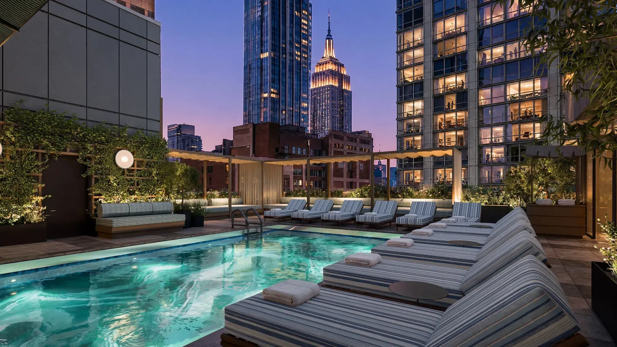 Rendering of a rooftop pool at dusk lined with striped loungers under a pergola, with lit towers and the Empire State Building behind